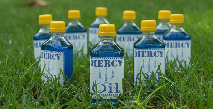 Bottle of 'Mercy Oil' with a yellow cap on a white surface, against a decorative background with text.